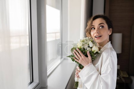 verblüffte junge Frau mit Verlobungsring am Finger in weißer Seidenrobe stehend und Brautstrauß haltend, während sie neben dem Fenster in der Hotelsuite nach oben schaut, besonderer Anlass, Braut am Hochzeitstag