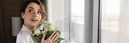 amazed woman with engagement ring on finger standing in white silk robe and holding bridal bouquet while looking up next to window in hotel suite, special occasion, bride on wedding day, banner 