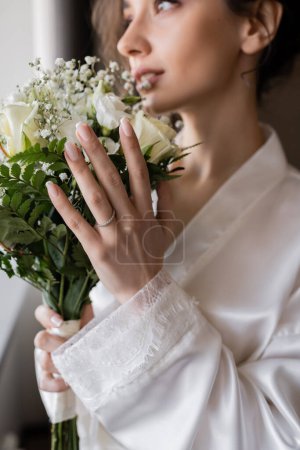 blurred bride with engagement ring on finger standing in white silk robe and holding bridal bouquet in modern hotel suite on wedding day, special occasion 