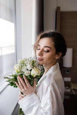 young woman with engagement ring on finger standing in white silk robe and looking at floral bridal bouquet next to window in hotel suite, special occasion, bride on wedding day