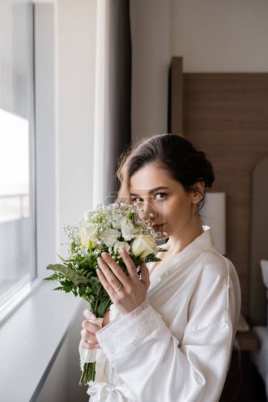 young bride with engagement ring on finger standing in white silk robe and smelling bridal bouquet next to window in hotel suite, special occasion, wedding day