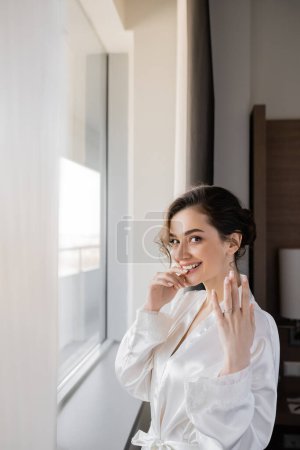 excited young bride with brunette hair in white silk robe showing engagement ring on finger and smiling next to window in hotel suit on wedding day, special occasion, hand near lips 