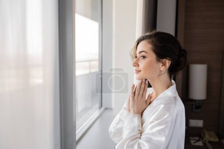 Photo for Side view of hopeful young woman with engagement ring on finger standing in white silk robe with praying hands and looking through window in hotel suite, special occasion, bride on wedding day - Royalty Free Image