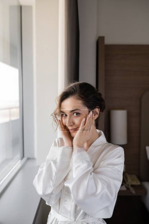 cheerful young bride with engagement ring on finger touching cheeks and standing in white silk robe next to window in hotel suite, special occasion, bride on wedding day