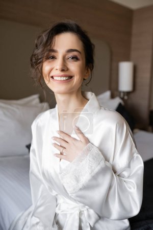 happy young bride with brunette hair and engagement ring on finger smiling while sitting in white silk robe and looking at camera in hotel room on wedding day, special occasion