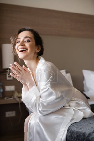 entzückende Braut mit brünetten Haaren, die lacht, während sie in einem weißen Seidenmantel auf einem bequemen Bett in einer modernen Hotelsuite am Hochzeitstag sitzt, besonderer Anlass 