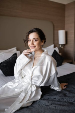 Photo for Pleased bride with brunette hair and bridal makeup touching chin and sitting in white silk robe on comfortable bed and looking at camera in hotel suite on wedding day - Royalty Free Image