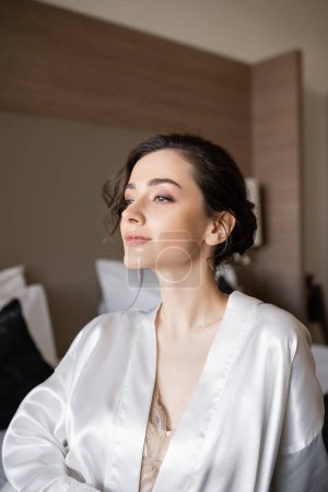 graceful young woman with brunette hair and bridal makeup sitting in white silk robe on comfortable bed and looking away in hotel suite on wedding day, special occasion 
