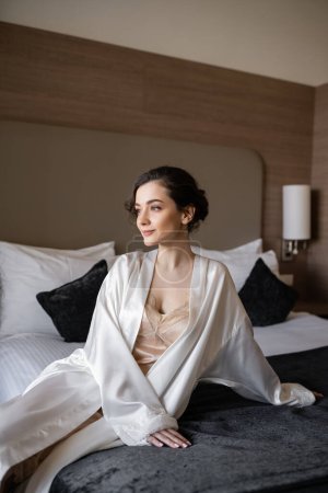 charming bride with brunette hair and bridal makeup sitting in white silk robe on comfortable bed and looking away in hotel suite before her wedding, special occasion 