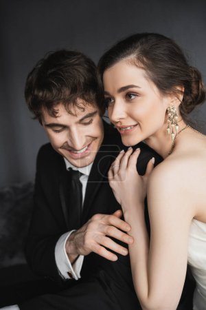 portrait of bride with brunette hair, elegant jewelry and white dress hugging shoulder of happy groom in classic formal wear with tie in modern hotel suite after wedding ceremony 