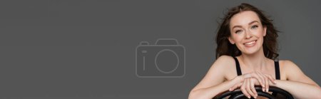 Téléchargez les photos : Jeune femme souriante et jolie aux cheveux bruns et au maquillage naturel regardant la caméra assise sur une chaise en bois isolée sur gris avec espace de copie, bannière - en image libre de droit