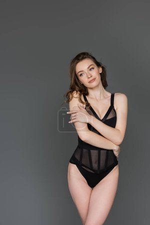 Pretty young brunette model with natural makeup and slim body posing in stylish black form-fitting bodysuit and looking away while standing isolated on grey  