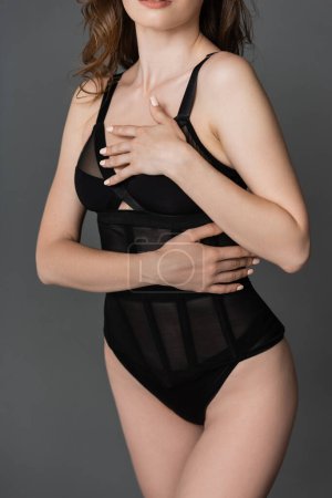 Ausgeschnittene Ansicht einer jungen und sinnlichen brünetten Frau mit schlankem Körper, die einen trendigen und formschlüssigen schwarzen Body trägt und die Brust berührt, während sie isoliert auf grau posiert  