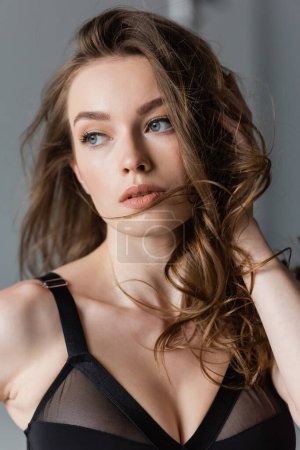 Portrait of young and confident model with brunette hair and natural makeup wearing black bodysuit and looking away while standing on grey background 