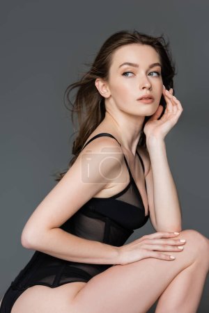 Portrait of sexy and fashionable brunette woman with natural makeup and modern hairstyle posing in black bodysuit and looking away while standing isolated on grey  