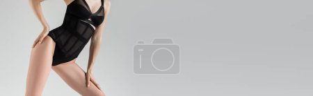 Foto de Vista recortada de la seductora modelo joven en elegante traje negro tocando la cadera y la pierna mientras está de pie y posando aislado en gris con espacio de copia, pancarta - Imagen libre de derechos