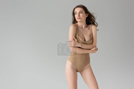 Trendy young woman with natural makeup and hairstyle touching arm and looking away while posing in sexy beige bodysuit isolated on grey with copy space