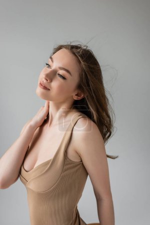 Portrait of pleased young brunette woman with everyday makeup touching neck and looking away while posing in sexy beige bodysuit isolated on grey  