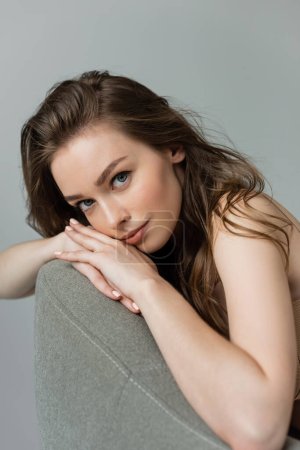 Portrait of pretty and young brunette woman with natural makeup and hairstyle looking at camera while relaxing on grey modern armchair isolated on grey  