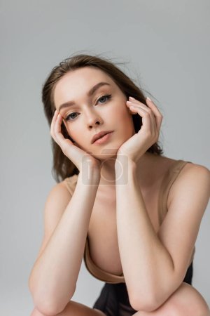 Portrait of young and seductive woman with brunette hair and natural makeup wearing bodysuit while touching face and looking at camera isolated on grey 