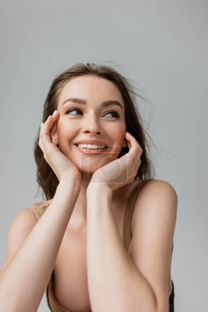 Portrait of cheerful and sexual young brunette woman with everyday makeup wearing bodysuit and looking away while posing isolated on grey, hands near face   
