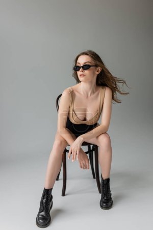 Full length of trendy young brunette model in sunglasses, beige bodysuit, black corset and boots posing while sitting on wooden chair on grey background 