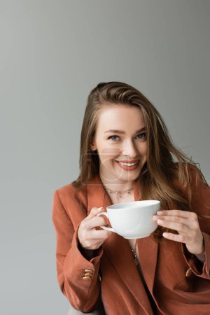 Portrait of smiling and fashionable young woman in terracotta jacket and necklaces holding cup of cappuccino and looking at camera isolated on grey  