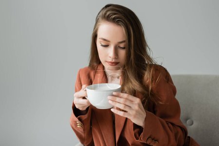 Trendige brünette junge Frau in Halskette und Terrakottamaske, die eine Tasse Cappuccino hält, während sie auf einem modernen Sessel sitzt, isoliert auf grau mit Kopierraum