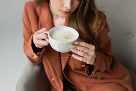 Ausgeschnittene Ansicht einer stilvollen jungen brünetten Frau im Terrakottablasser, die eine Tasse Cappuccino hält, während sie auf einem verschwommenen und bequemen Sessel sitzt, isoliert auf grau  