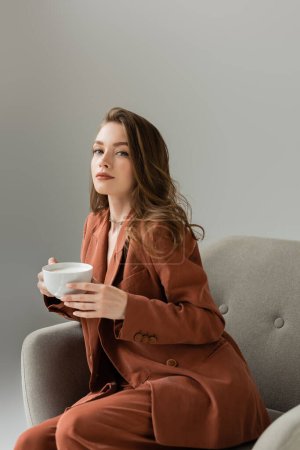 Fashionable young woman in terracotta suit and necklace looking at camera while holding cup of cappuccino and sitting on modern armchair isolated on grey  