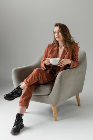 longitud completa de la mujer joven con el pelo largo y collar con traje de moda de terracota con chaqueta y pantalones y la celebración de la taza de café mientras está sentado en el sillón sobre fondo gris en el estudio 