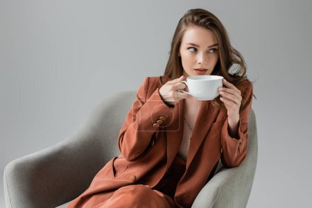 brünette junge Frau mit langen Haaren und Halskette in Terrakotta und trendigem Anzug mit Blazer und Hose und Kaffee trinken, während sie in einem bequemen Sessel auf grauem Hintergrund im Studio sitzt 