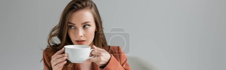 brunette young woman with long hair and necklace wearing trendy suit with blazer and drinking coffee while looking away on grey background in studio, sophisticated lady, coffee break, banner