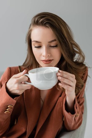 brünette junge Frau mit langen Haaren im trendigen Anzug mit Blazer und Kaffeetasse im bequemen Sessel auf grauem Hintergrund im Studio sitzend, Work-Life-Balance, Terrakotta 