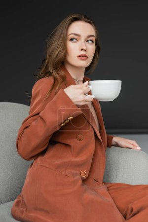 young woman with long hair wearing brown and trendy suit with blazer and pants and holding cup of coffee while sitting in comfortable armchair on grey background, looking away, work-life balance