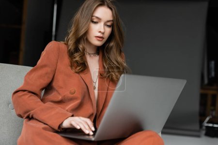brunette young woman with long hair and necklace wearing terracotta stylish suit with blazer and pants using laptop while sitting in comfortable armchair on grey background, freelancer, remote work 