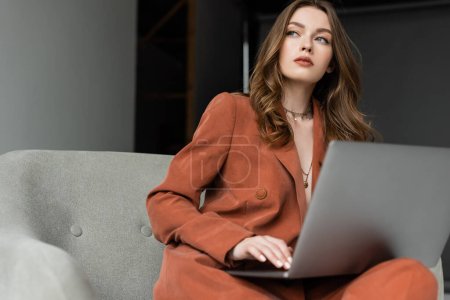 pensive young woman with long hair and necklace wearing trendy suit with blazer and pants and using laptop while sitting in comfortable armchair on grey background, freelancer, remote work 