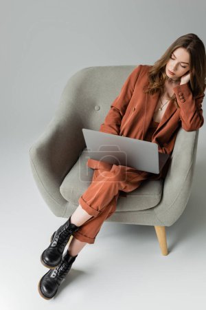 Ansicht der brünetten jungen Frau mit langen Haaren und Halskette im trendigen Anzug mit Blazer und Hose und Laptop im Sessel auf grauem Hintergrund, freischaffend