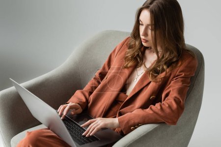 brünette junge Frau mit langen Haaren und Halskette, die einen trendigen Terrakotta-Anzug mit Blazer und Hose trägt und auf dem Laptop tippt, während sie in einem bequemen Sessel auf grauem Hintergrund sitzt, Fernarbeit