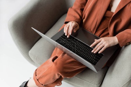 vista superior de la mujer joven que usa traje de moda con chaqueta y pantalones usando el ordenador portátil mientras está sentado en un cómodo sillón sobre fondo gris, freelancer, trabajo remoto, tiro recortado 