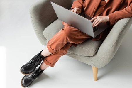 top view of young woman wearing terracotta trendy suit with blazer and pants with boots using laptop while sitting in comfortable armchair on grey background, freelancer, remote work, cropped shot