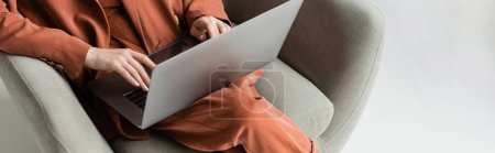 vista superior de la mujer joven que usa traje de moda de terracota con chaqueta y pantalones usando el ordenador portátil mientras está sentado en cómodo sillón sobre fondo gris, freelancer, trabajo remoto, tiro recortado, pancarta
