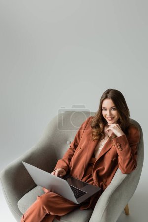 sonriente mujer joven con el pelo largo y collar de terracota traje de moda con chaqueta y pantalones y el uso de ordenador portátil mientras está sentado en el sillón sobre fondo gris, freelancer, mira a la cámara 