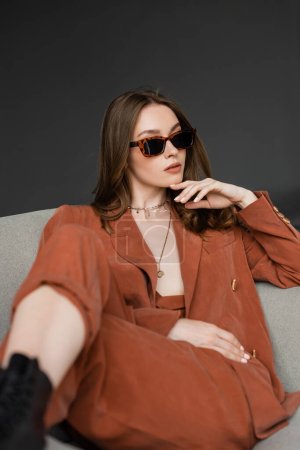 stylische junge Frau mit langen Haaren trägt Terrakotta-Anzug mit Blazer und Hose und posiert mit trendiger Sonnenbrille, während sie im bequemen Sessel auf grauem Hintergrund sitzt, modisches Modell, schaut weg