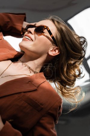 Foto de Vista superior de la joven feliz con el pelo largo y maquillaje natural con terracota y traje de moda con chaqueta y collar de oro mientras posan en gafas de sol sobre fondo gris, modelo hermoso - Imagen libre de derechos