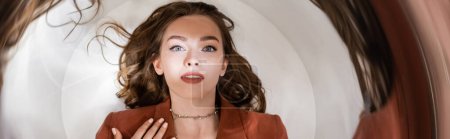 Photo for Circular shot, top view of alluring young woman with brunette hair in blazer and golden necklace lying and looking at camera on grey background, blurred, wide-angle, distorted view, banner - Royalty Free Image