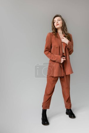 Photo for Full length of stylish young woman with brunette hair and necklace posing in terracotta and trendy suit with blazer and pants, black boots while looking away on grey background in studio, trendy model - Royalty Free Image