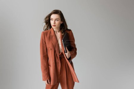 brunette young woman with long hair and necklace wearing terracotta trendy suit with blazer and pants, holding laptop while standing on grey background, freelancer, remote work, looking away