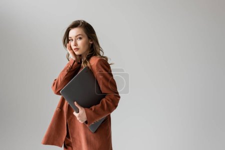 Photo for Pretty young woman with brunette hair and necklace wearing terracotta and trendy suit with blazer, looking at camera and holding laptop while standing on grey background, freelancer, remote work - Royalty Free Image