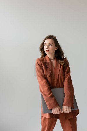 young woman with brunette hair and necklace wearing terracotta and trendy suit with blazer and pants, looking away and holding laptop while standing on grey background, freelancer, remote work 